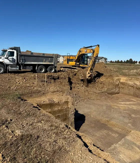 Yellow excavator removing soil for a building foundation with a dump truck parked nearby on a construction lot